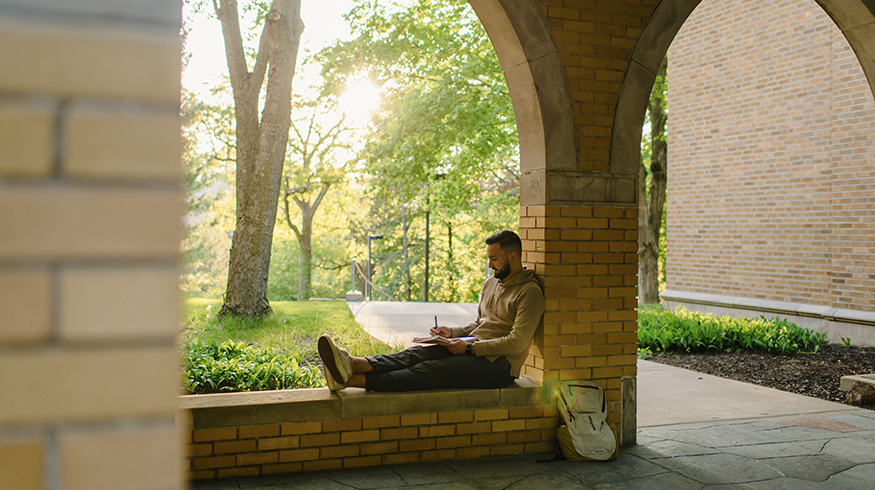 Student sits in arches and works in notebook.