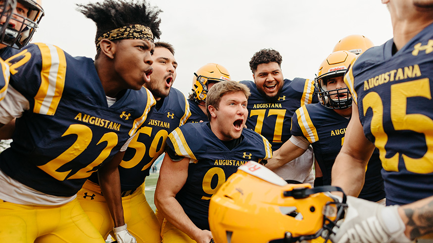 A group of football players shouting in a huddle on Augustana's field