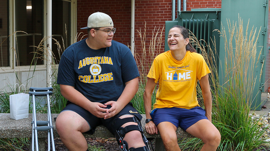 President Andrea Talentino laughs next to a male student, both sitting outside a residence hall