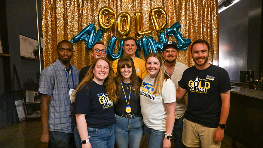 Young alumni pose in front of a yellow cloth backdrop with balloons that spell out "GOLD ALUMNI"
